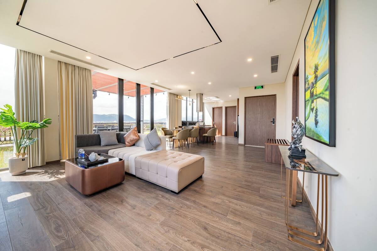 A spacious hotel lobby with a modern design, featuring a seating area with a sofa and ottoman, a dining area, and large windows offering a view of the outdoors.