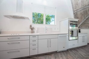 A modern kitchen with white cabinetry, a stainless steel oven, and a window with a view of trees. - Sumner Lake House 2 Bedrooms (Clermont)