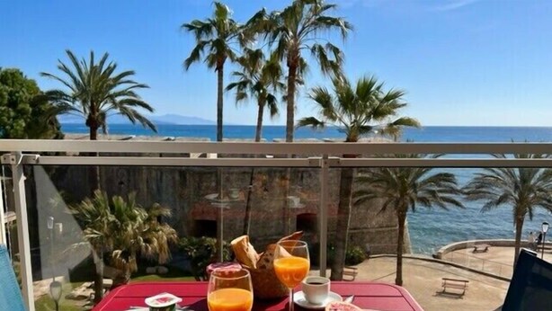 Une table dressée avec des produits pour le petit-déjeuner, notamment des croissants, des fruits et des boissons, offrant une vue sur la côte et les palmiers.