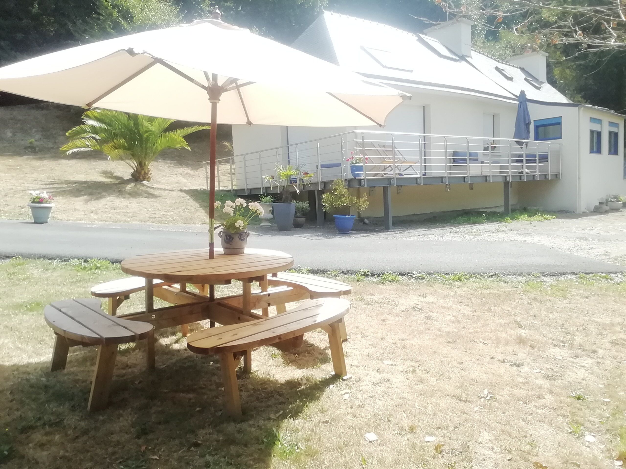 A wooden outdoor table with a beige umbrella, surrounded by potted plants and a house with a balcony in the background.