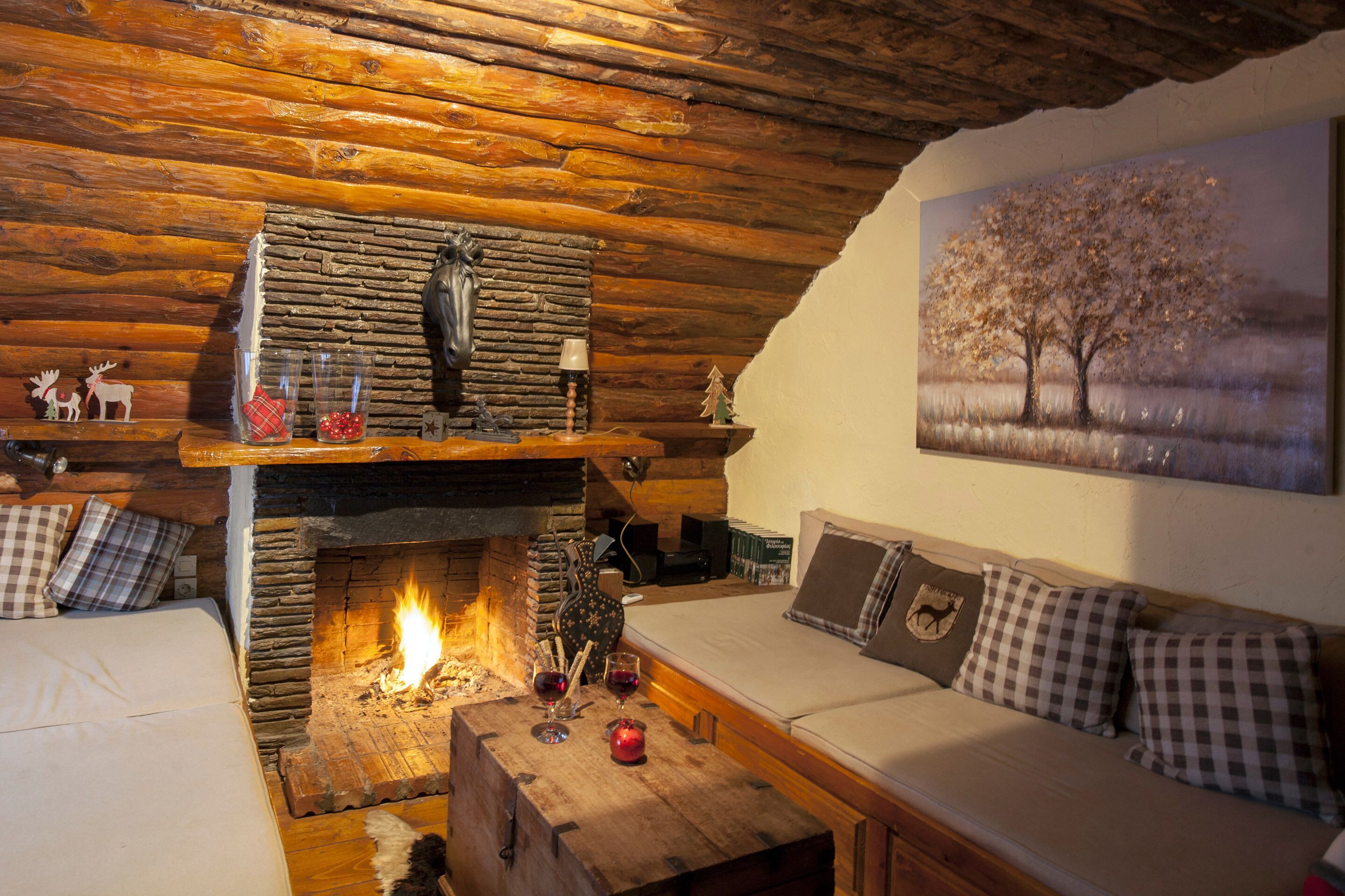 Interior acolhedor de uma cabana de toras, com uma lareira de pedra, um sofá com almofadas xadrez e um quadro de uma árvore na parede.