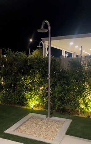 A modern outdoor shower with a metal frame, surrounded by a square gravel area and lush green bushes. - A modern vacation home in Desert Hot Springs, built in 2020 with 4 bd, 3 baths (Desert Hot Springs)