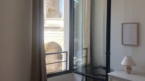 A bedroom with a bed, a dresser, a lamp, and a window with a view of an archway. - Opposite Les Arenes and La Major (Arles)