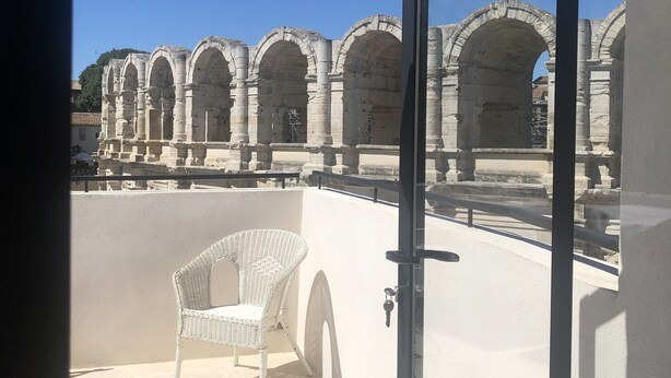 A balcony with a white wicker chair, a glass door, and a view of ancient stone arches. - Opposite Les Arenes and La Major (Arles)