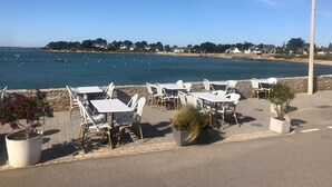 A seaside patio with tables and chairs, a stone wall, and a view of the sea and distant buildings.