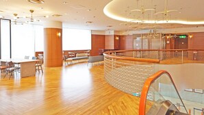 A spacious lobby with a wooden floor, a seating area with chairs and tables, a glass railing, and an escalator. - Chisun Hotel Yokohama Isezakicho (Yokohama)