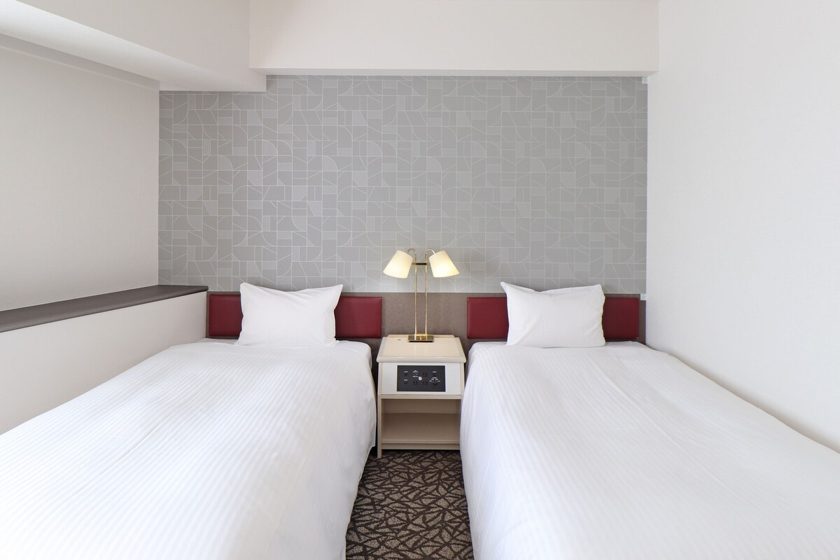 Habitación de hotel con dos camas individuales, una mesita de noche con lámpara y una alfombra estampada.