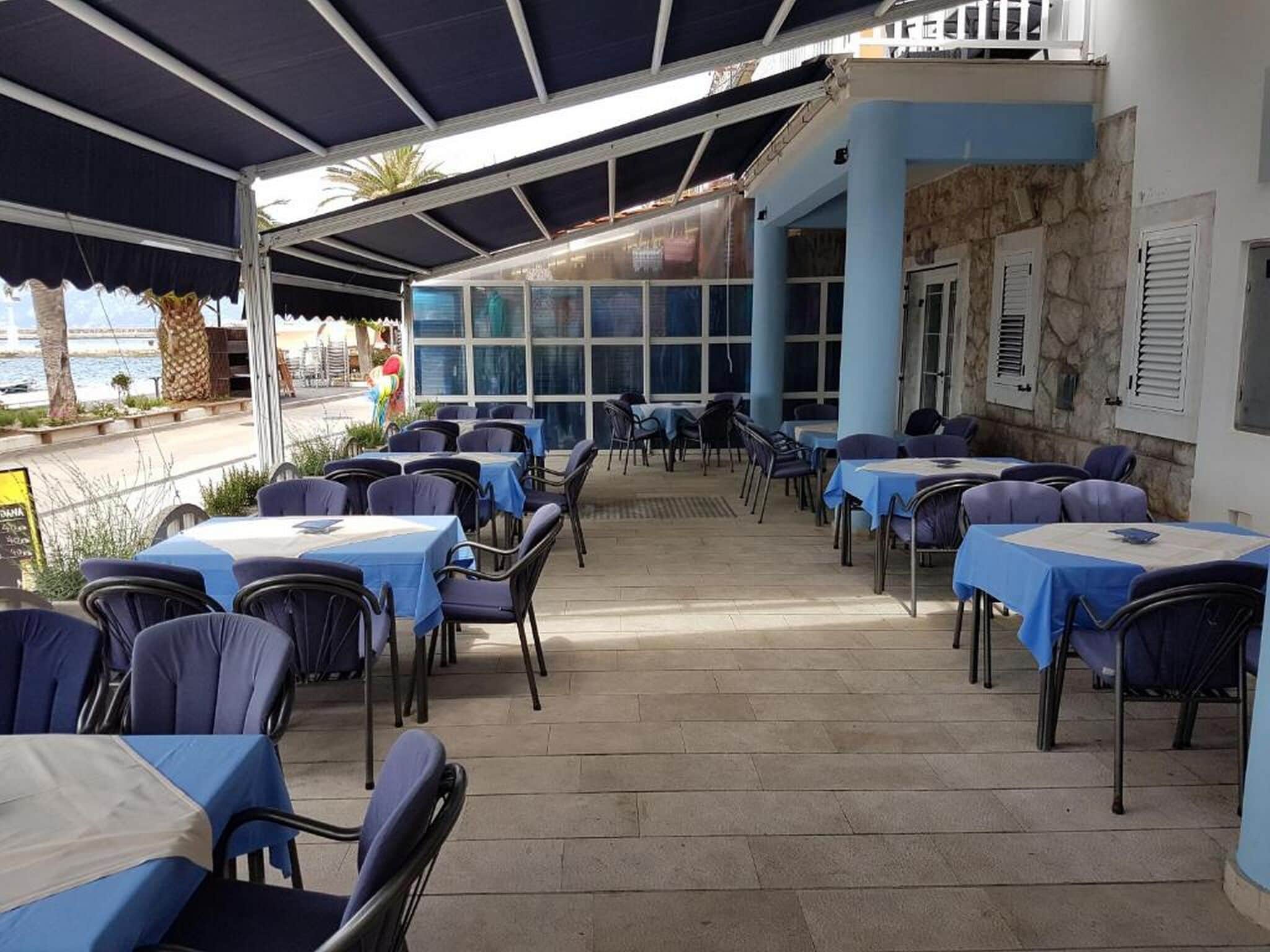 Ein Straßencafé mit Tischen und Stühlen, eine Steinmauer und Blick auf das Meer.