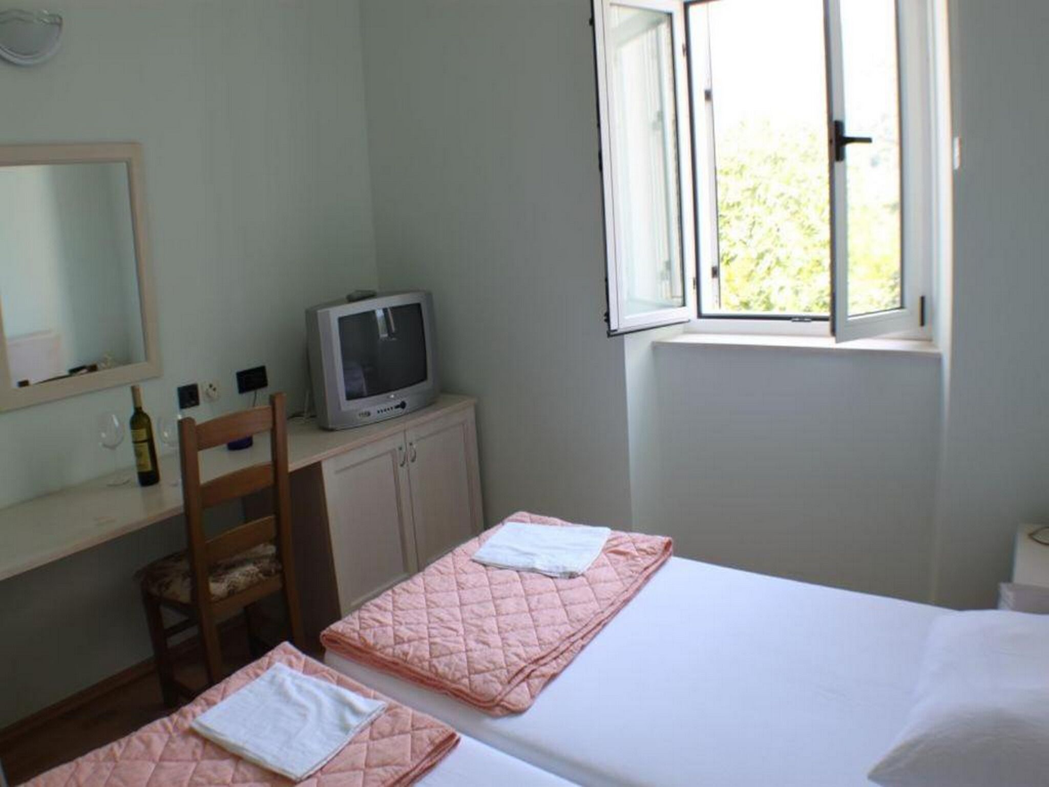 Quarto com duas camas, uma TV sobre um móvel, uma cadeira e uma janela com vista para áreas verdes.