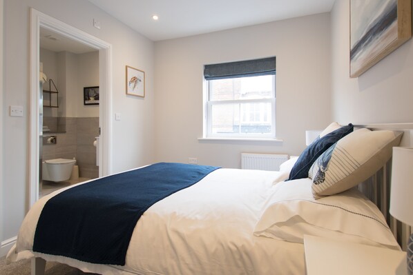 Une chambre à coucher avec un lit, une fenêtre, un radiateur et une salle de bain visible à travers une porte ouverte.
