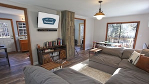 A living room with a large gray sectional sofa, a wooden coffee table, and a bookshelf. There is a large window with a view of trees and a deck. - Dream Stratton Forest Cabin w/Hot Tub & Fast WiFi (Stratton)