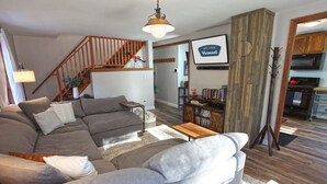 A living room with a grey sectional sofa, a wooden staircase, a bookshelf, and a TV displaying "We Love Vermont". - Dream Stratton Forest Cabin w/Hot Tub & Fast WiFi (Stratton)