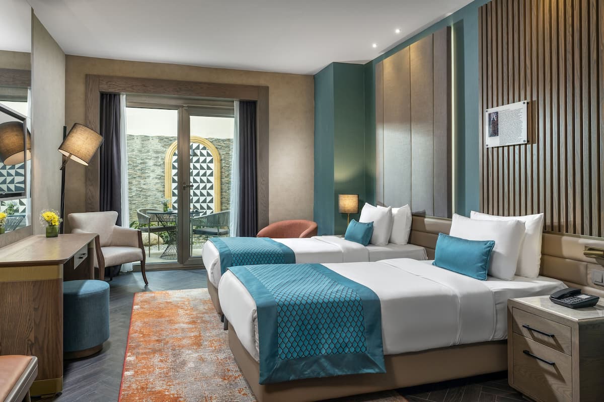 Une chambre d’hôtel avec deux lits, un bureau, une chaise, une petite table avec des fleurs et une vue sur l’extérieur.