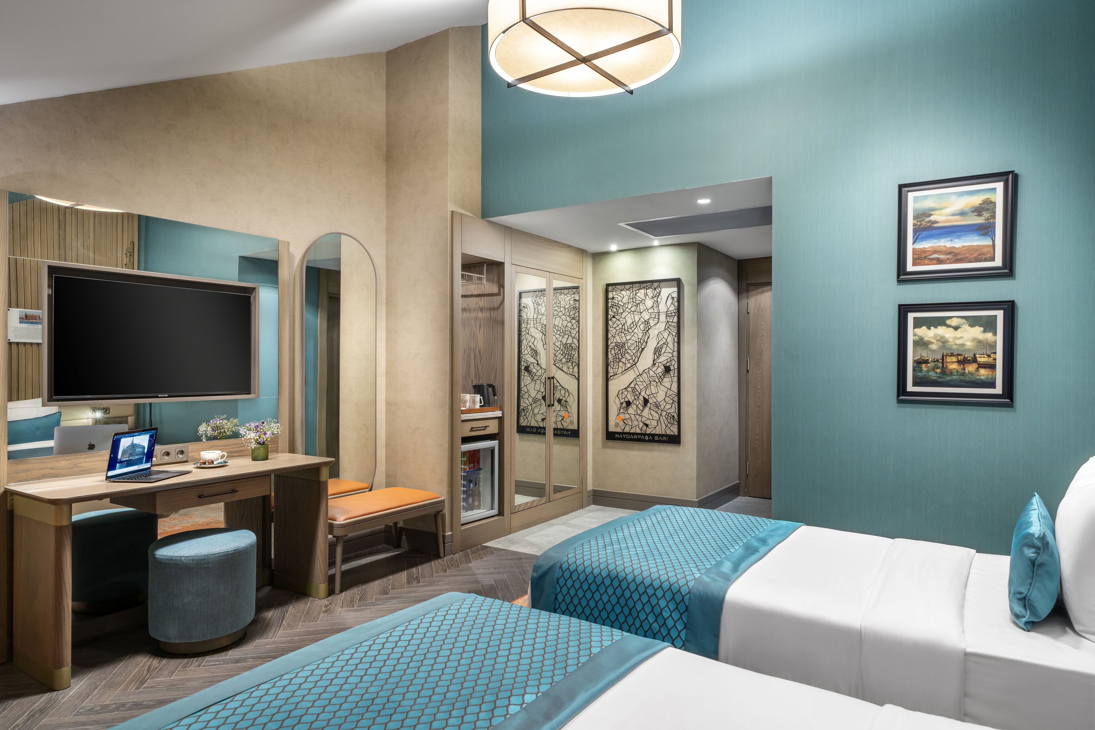 Une chambre d’hôtel moderne dotée d’un grand téléviseur à écran plat, d’un bureau avec un ordinateur portable, d’un coin salon confortable, de deux lits avec des têtes de lit assorties et d’œuvres d’art accrochées aux murs.