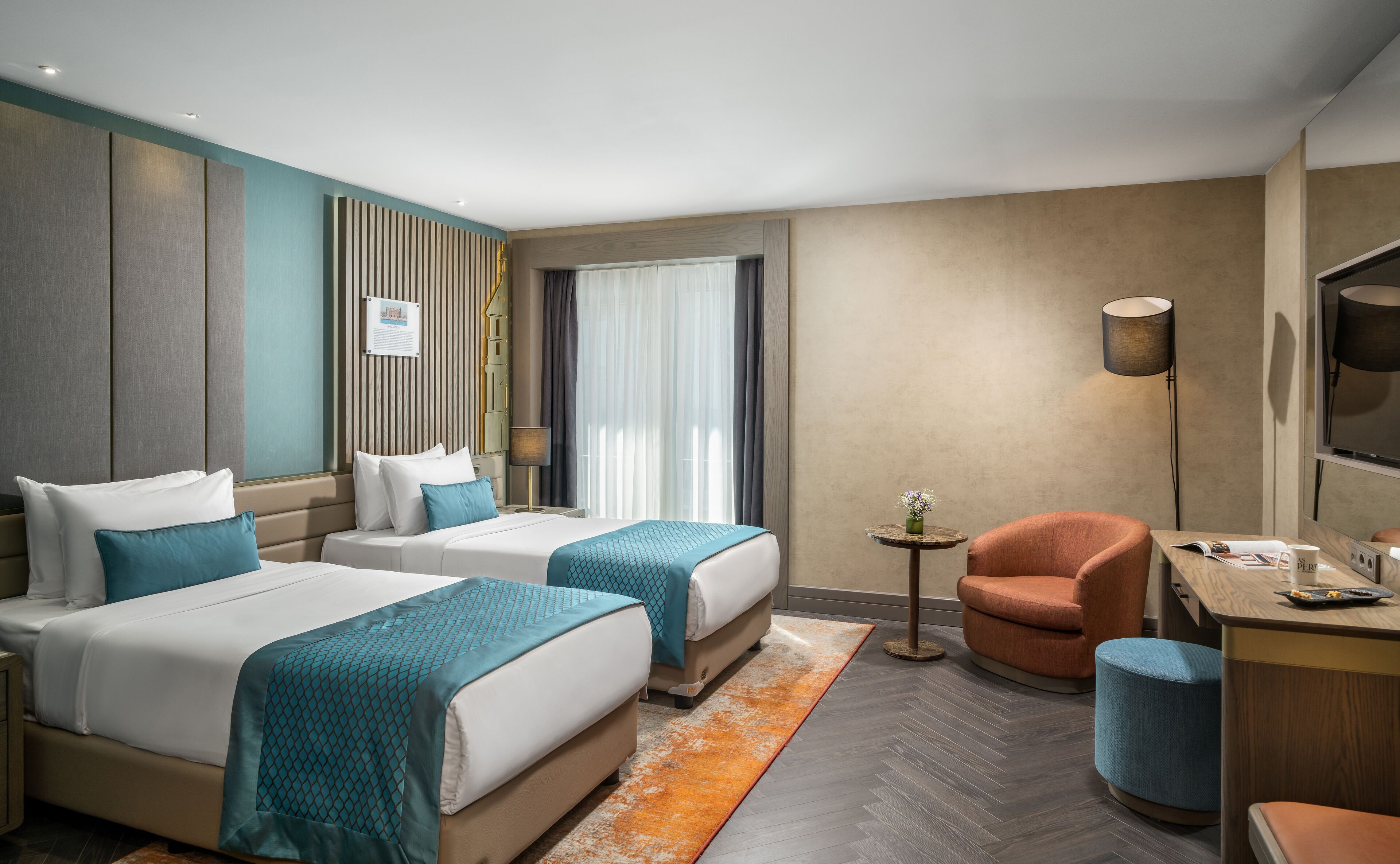 A hotel room with two beds, a desk, a chair, a small table, and a lamp.