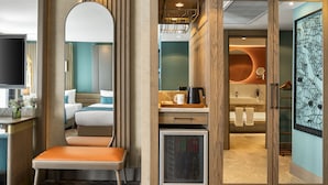 Une chambre d’hôtel moderne équipée d’un lit, d’un coin salon, d’un minibar et d’une salle de bain séparée par une paroi en verre.