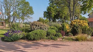 Une allée de gravier mène à une maison entourée de divers jardins aménagés, où l’on trouve différentes espèces de plantes et d’arbres.