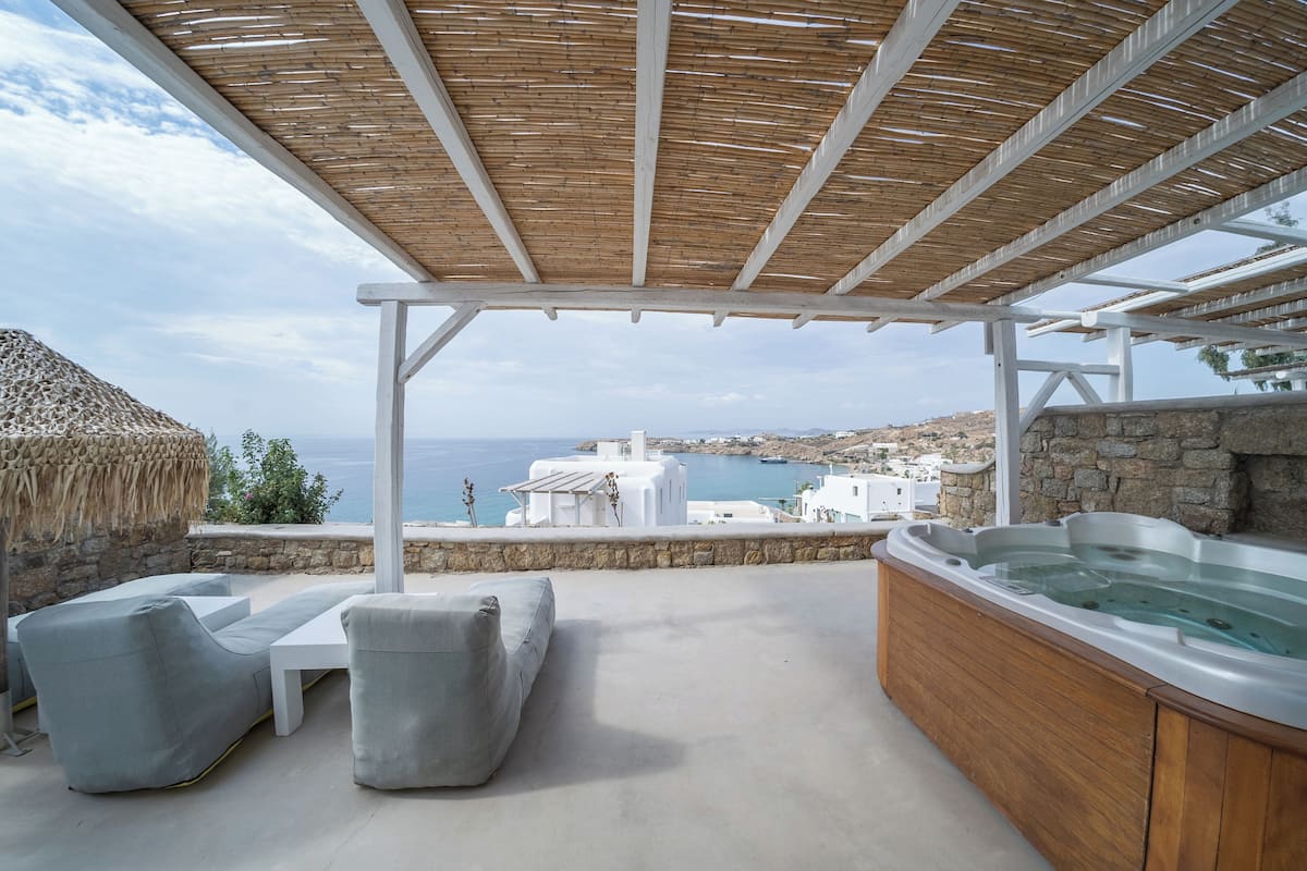 A covered outdoor seating area with a hot tub, white furniture, and a view of the sea and buildings.