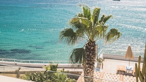 A seaside view with a palm tree, a pool area, and boats on the water. - New Paradise Beach Resort- Adults Only (Mykonos)