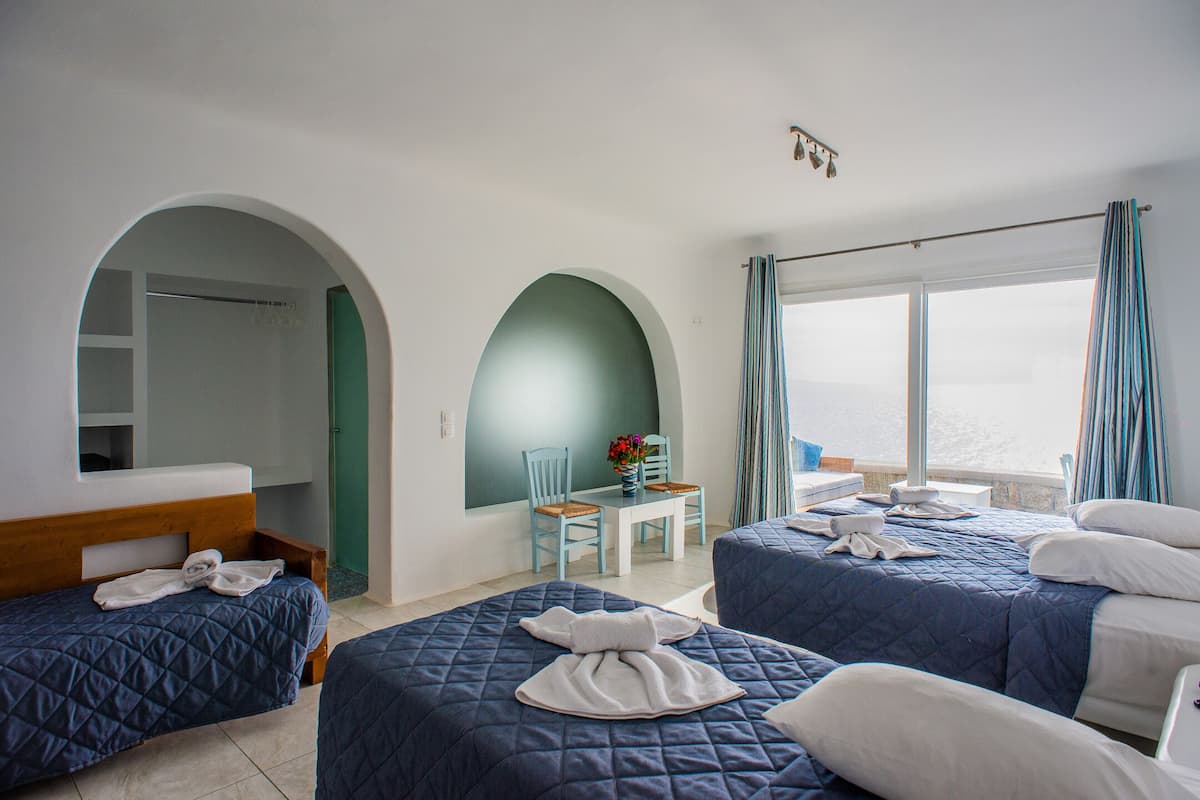 A hotel room with two beds, a small table with chairs, a view of the sea, and a balcony with curtains.