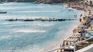 A coastal beach with boats, a sandy shore, and buildings in the background. - New Paradise Beach Resort- Adults Only (Mykonos)