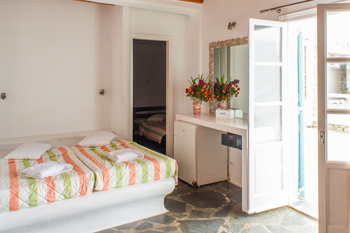 A bedroom with a stone floor, wooden ceiling beams, a bed with a striped blanket, a desk with a mirror, and a door leading outside.