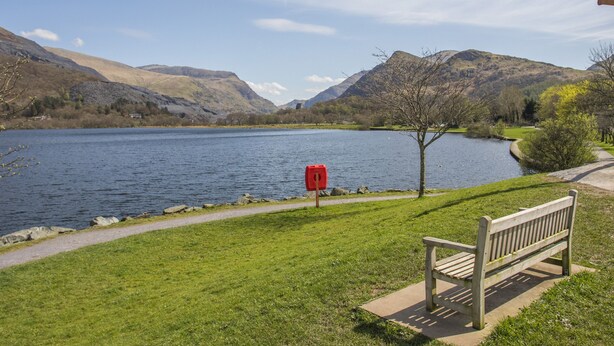 A bench by a lake with a red mailbox and a path leading to a grassy area. - 9 Ty Du Road (Caernarfon)