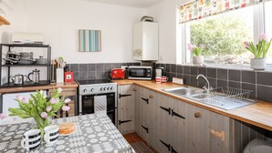 A modern kitchen with a stove, microwave, and sink. There is a window with plants on the windowsill and a shelf with kitchen utensils. - 9 Ty Du Road (Caernarfon)