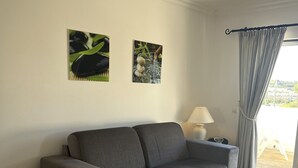Ett vardagsrum med en grå soffa, ett litet bord med en vävd bordsduk, två inramade tavlor på väggen, en lampa på ett sidobord och en utsikt genom ett fönster med gardiner.