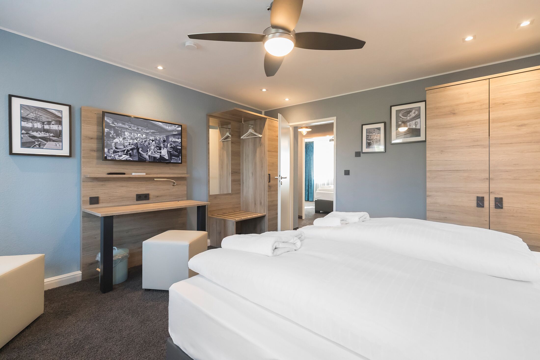 A modern hotel room with a large bed, a desk with a flat-screen TV, a ceiling fan, and a wardrobe.