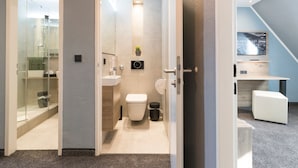 Un baño moderno con inodoro, lavamanos y ducha. Hay una planta pequeña en la pared y un televisor en la habitación contigua.