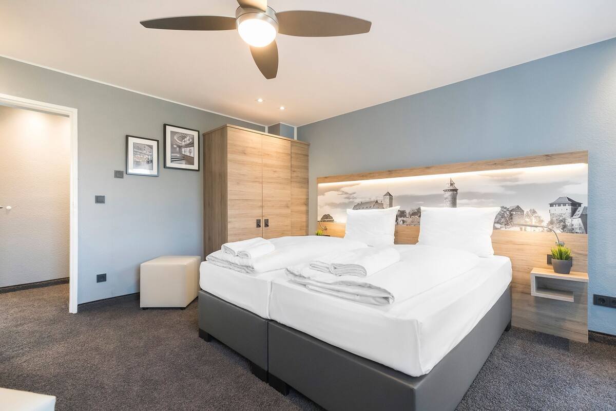 A modern bedroom with a large bed, a ceiling fan, a nightstand, a small potted plant, and a wall-mounted shelf.