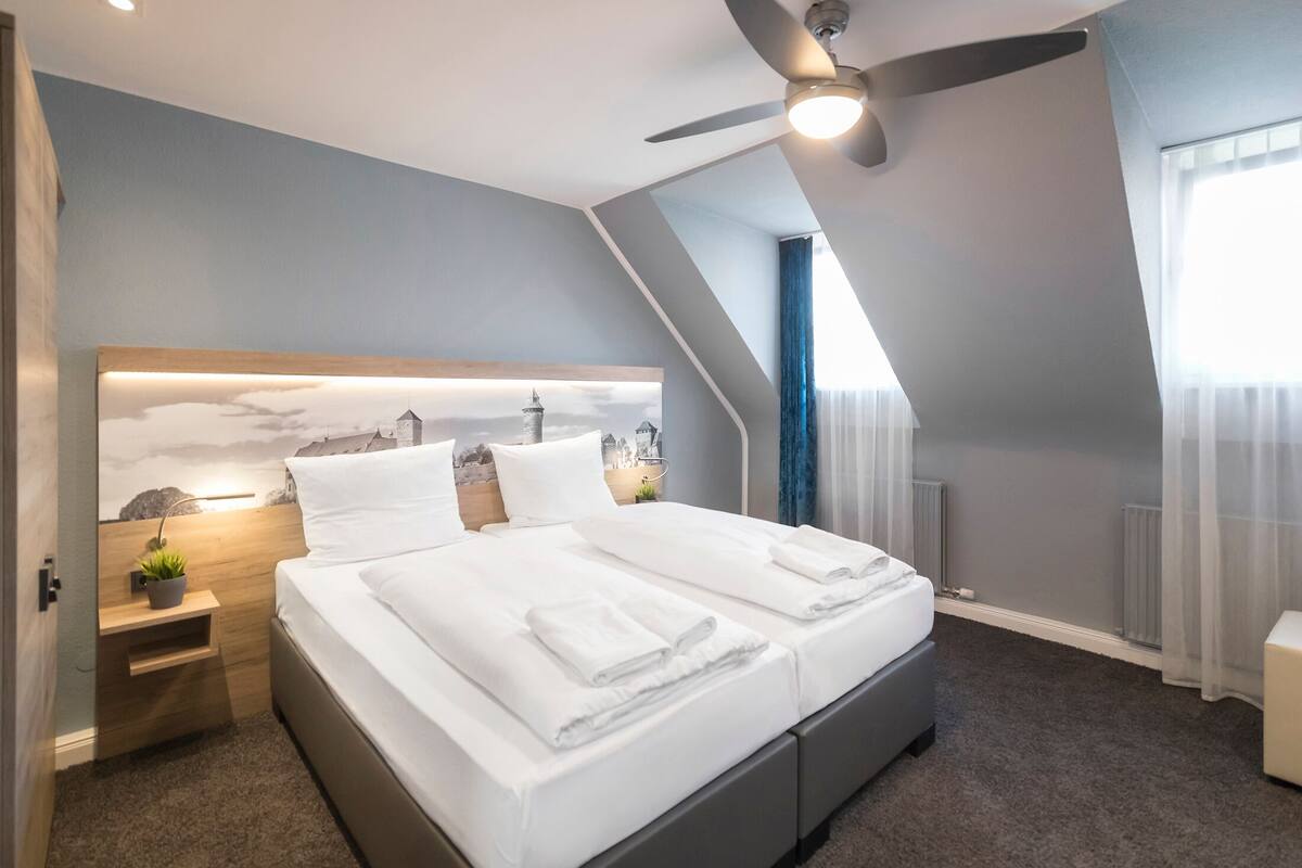 A modern hotel room with a large bed, a ceiling fan, a nightstand with a plant, and a cityscape mural on the wall.