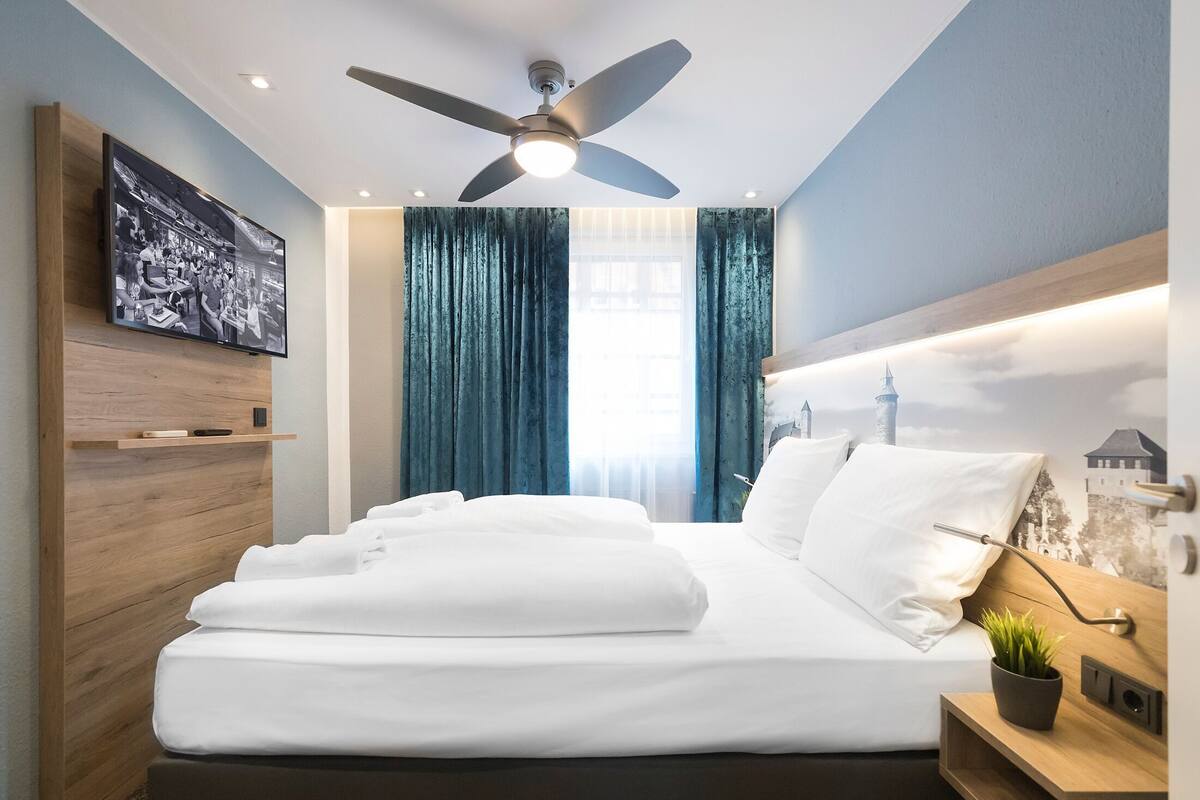 A hotel room with a bed, a ceiling fan, a mounted TV, a window with curtains, and a wall-mounted shelf.
