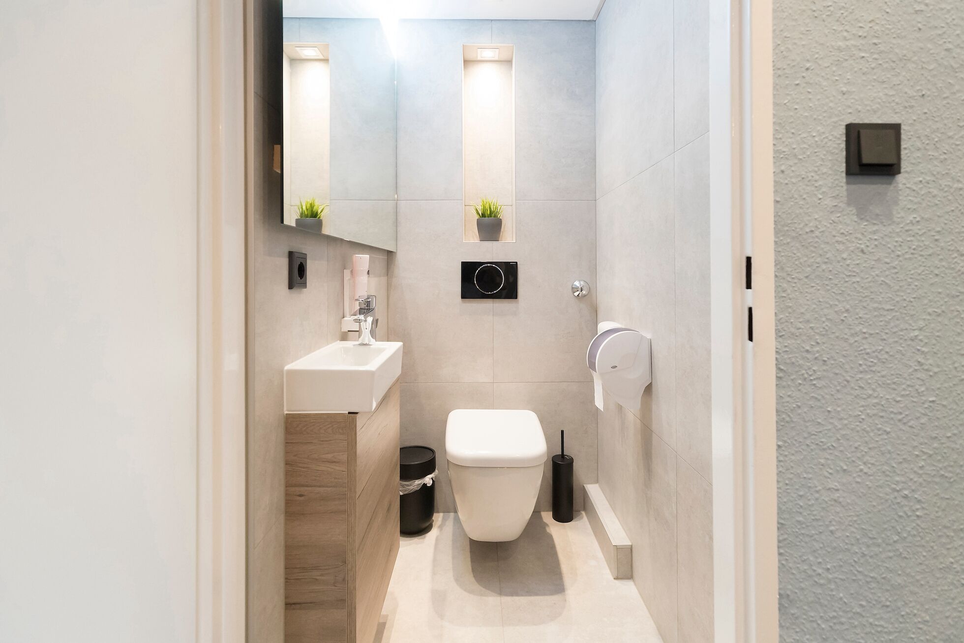 Ein modernes Badezimmer mit einem weißen Waschbecken, einer Toilette und einem Spiegel über dem Waschbecken. Es befinden sich zwei Wandleuchten und ein kleiner Topfpflanze an der Wand.