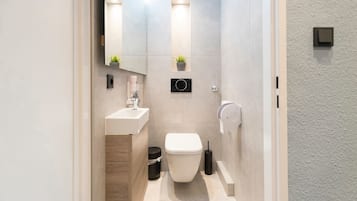 Ein modernes Badezimmer mit einem weißen Waschbecken, einer Toilette und einem Spiegel über dem Waschbecken. Es befinden sich zwei Wandleuchten und ein kleiner Topfpflanze an der Wand.
