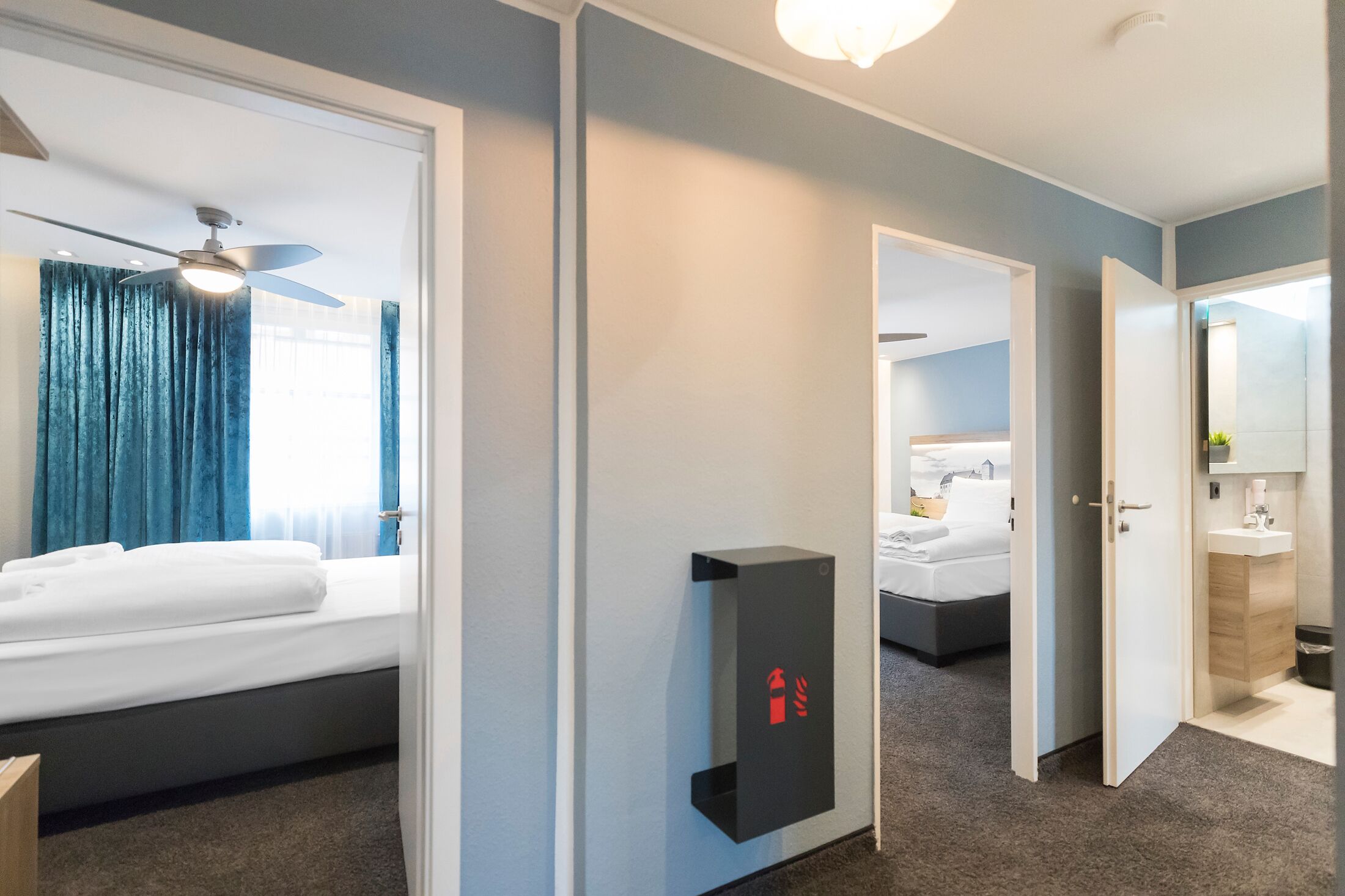 Une chambre d’hôtel comprenant un lit, un ventilateur de plafond, un détecteur d’incendie fixé au mur, et une salle de bain avec un lavabo et un miroir.