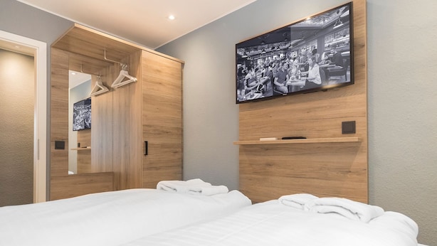Habitación de hotel con un armario de madera, una cama con ropa de cama blanca y un televisor en la pared mostrando una imagen en blanco y negro.
