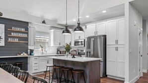 Una cucina moderna con mobili bianchi, una grande isola con sgabelli, illuminazione a sospensione e un cartello appeso al muro.