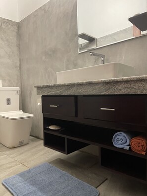 Ein modernes Badezimmer mit grauer Marmorwand, einer weißen Toilette, einem dunklen Holzwaschtisch mit Spiegel und einer blauen Badematte.