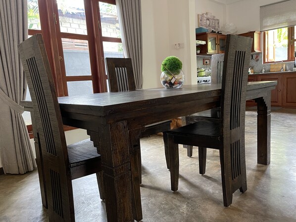 A dining area with a wooden table and chairs, a vase with a plant, and a kitchen in the background.