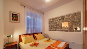 Hotelkamer met een bed, nachtkastje, lamp, raam met dunne gordijnen, stenen muur met een verlichtingsarmatuur en een lijst met een afbeelding.