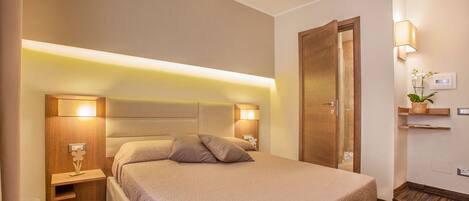 Habitación de hotel con una cama grande, lámparas de noche, cabecero de madera, mesita de noche, estante en la pared con una planta y una puerta que da a otra habitación.