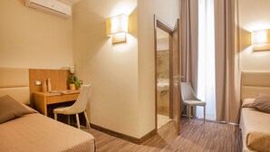 Habitación de hotel con dos camas, un escritorio y una silla, una mesita con un jarrón y una planta, y un baño visible a través de una puerta de vidrio.