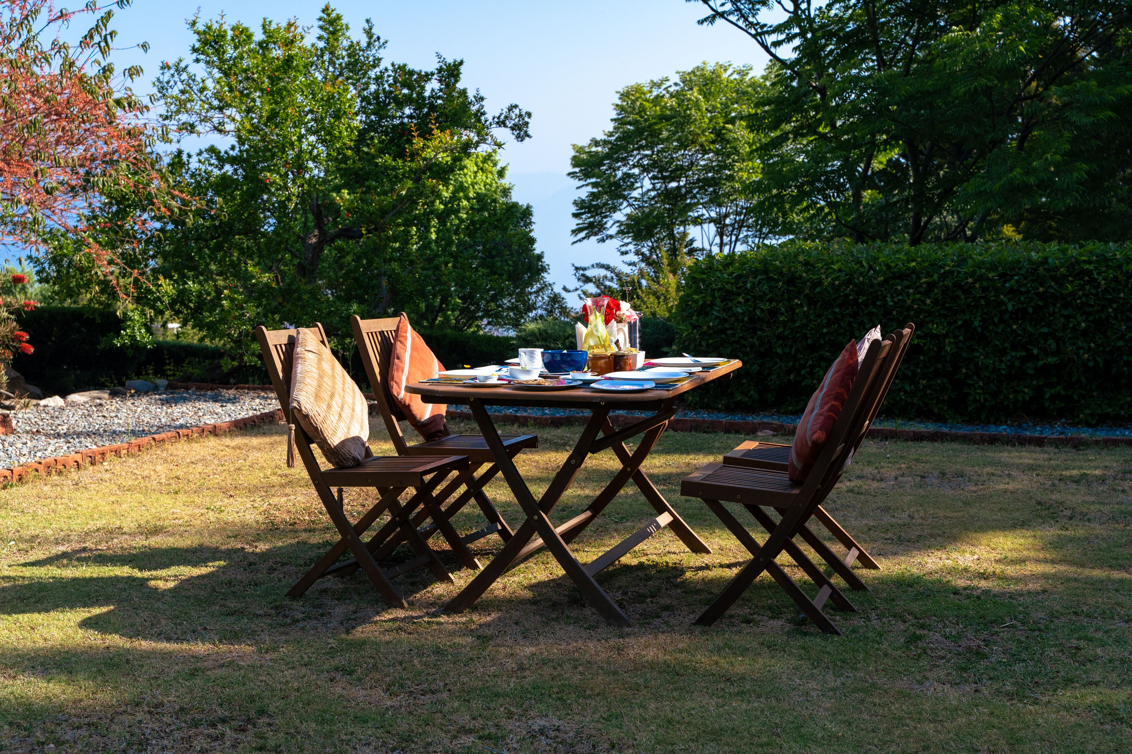 An outdoor dining area with a table and four chairs, surrounded by a garden with trees and a hedge.