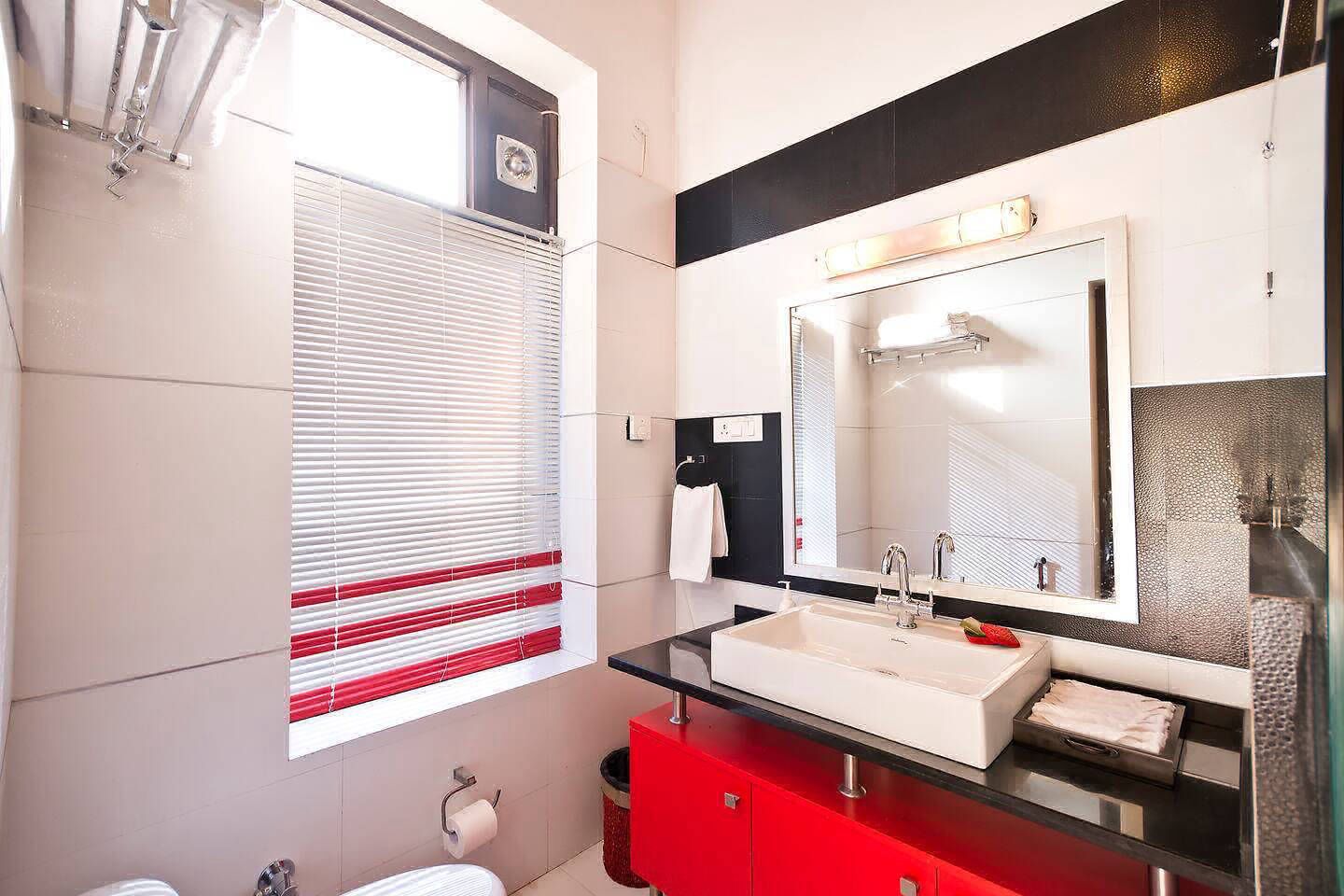 A modern bathroom with a large mirror, white sink, red cabinet, and a window with blinds.