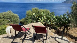 Ein Küstenblick mit zwei roten Stühlen, die auf das Meer ausgerichtet sind, umgeben von Grünpflanzen und einem Steintisch.