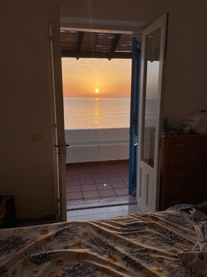 A room with a bed, a view of the sea at sunset, and a tiled floor.