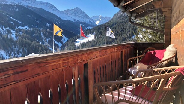Un balcon en bois, agrémenté de chaises en osier, qui offre une vue sur un paysage montagneux parsemé de drapeaux.