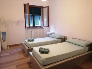 Une chambre avec deux lits simples, une fenêtre avec des stores, un radiateur et une petite table blanche.